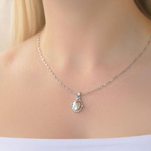 May include: A silver necklace with a teardrop-shaped pendant. The pendant has a central pearl-like stone and a small blue accent stone. The necklace is worn on a person wearing a white top.