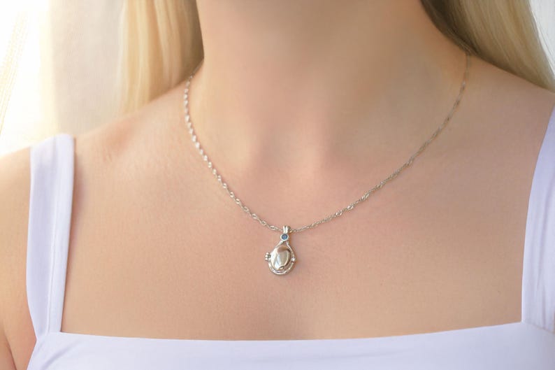 May include: A silver necklace with a teardrop-shaped pendant. The pendant has a central pearl-like stone and a small blue accent stone. The necklace is worn on a person wearing a white top.