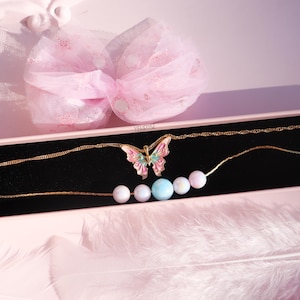 May include: A gold chain necklace with a pink and green butterfly pendant and a strand of pastel colored beads. The necklace is displayed in a pink box with a white tulle bow.