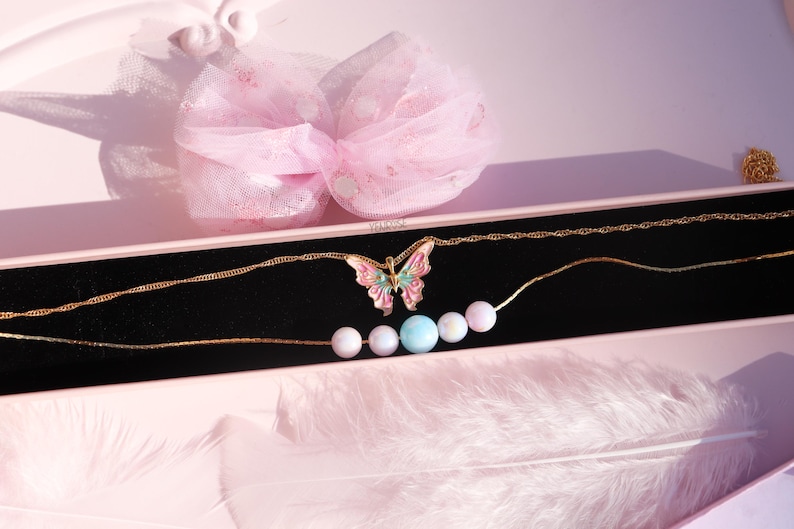 May include: A gold chain necklace with a pink and green butterfly pendant and a strand of pastel colored beads. The necklace is displayed in a pink box with a white tulle bow.