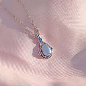 May include: A silver-toned pendant necklace with a teardrop-shaped pendant. The pendant has a light-colored oval stone and a small blue accent stone. The necklace is displayed on a pink, silky fabric.