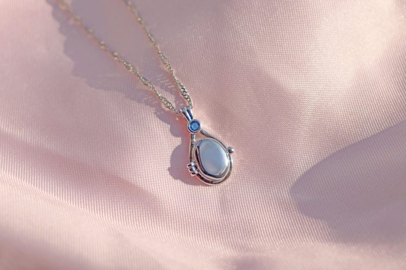 May include: A silver-toned pendant necklace with a teardrop-shaped pendant. The pendant has a light-colored oval stone and a small blue accent stone. The necklace is displayed on a pink, silky fabric.