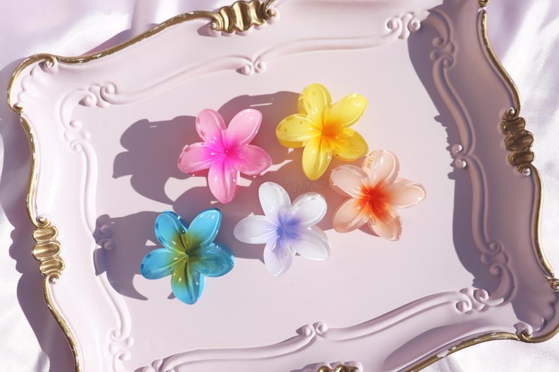 May include: Six colorful plastic flower hair clips on a pink tray. The clips are in shades of pink, yellow, blue, orange, and white.