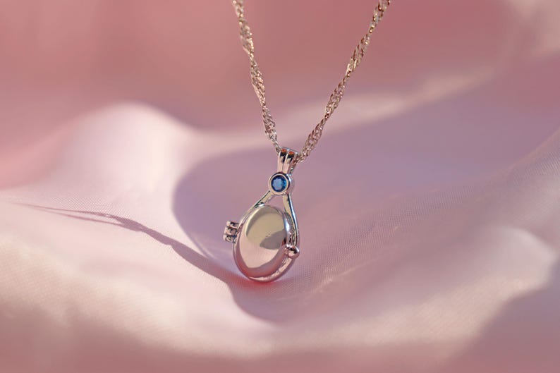 May include: A silver-toned necklace with a teardrop-shaped pendant. The pendant has a small blue gemstone at the top and a hinged design. The necklace hangs against a soft pink background.