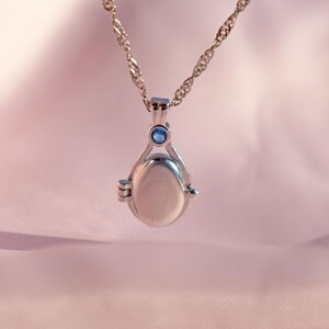 May include: Silver-toned locket necklace with a delicate chain. The pendant has an oval shape with a small blue gemstone at the top. The locket is suspended against a soft pink background.