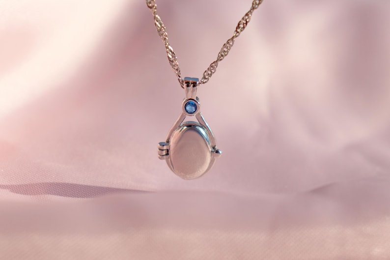 May include: Silver-toned locket necklace with a delicate chain. The pendant has an oval shape with a small blue gemstone at the top. The locket is suspended against a soft pink background.