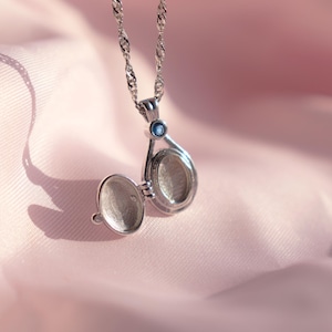 May include: Silver locket necklace with an open teardrop-shaped pendant. The locket is on a delicate chain and has a small blue gemstone. The background is a soft pink fabric, creating a romantic and elegant feel.