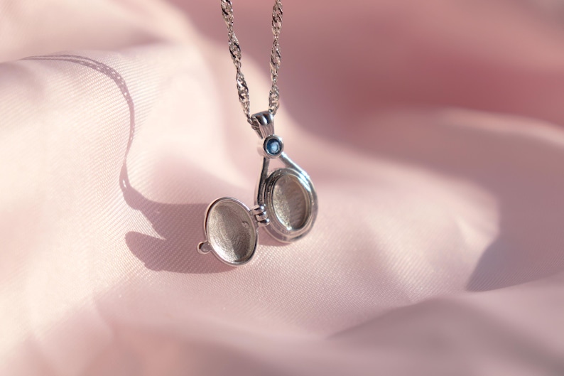 May include: Silver locket necklace with an open teardrop-shaped pendant. The locket is on a delicate chain and has a small blue gemstone. The background is a soft pink fabric, creating a romantic and elegant feel.