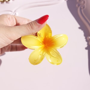 May include: A yellow resin flower with five petals and a center that is a darker yellow. The flower is held in a hand.