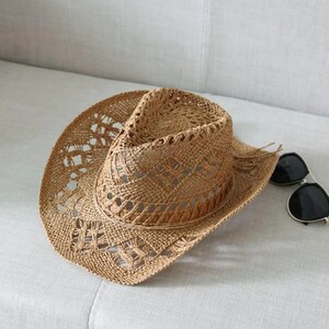 Puede incluir: Sombrero de vaquero de paja tejida de color marr&oacute;n claro, con ala ancha y patrones decorativos calados. El sombrero tiene una banda marr&oacute;n alrededor de la corona. Gafas de sol negras sobre el ala.