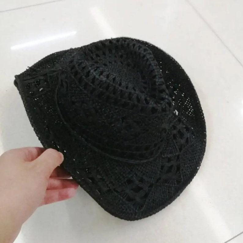 Puede incluir: Un sombrero de sol negro de tejido abierto con ala ancha. El sombrero est&aacute; hecho de un material tejido y tiene un aspecto ligeramente desgastado. El sombrero est&aacute; sostenido por una mano.