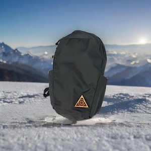 Könnte beinhalten: Ein schwarzer Rucksack mit einem dreieckigen Holz-Emblem mit einem kreisförmigen Design. Der Rucksack befindet sich vor einer schneebedeckten Berglandschaft mit einer hellen Sonne im Hintergrund. Der Rucksack hat ein stromlinienförmiges Design.