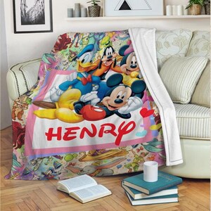 May include: A colorful Disney-themed blanket featuring Mickey Mouse, Donald Duck, and Goofy. The blanket has a pink border with the name "HENRY" in red script. The background is filled with various Disney characters.