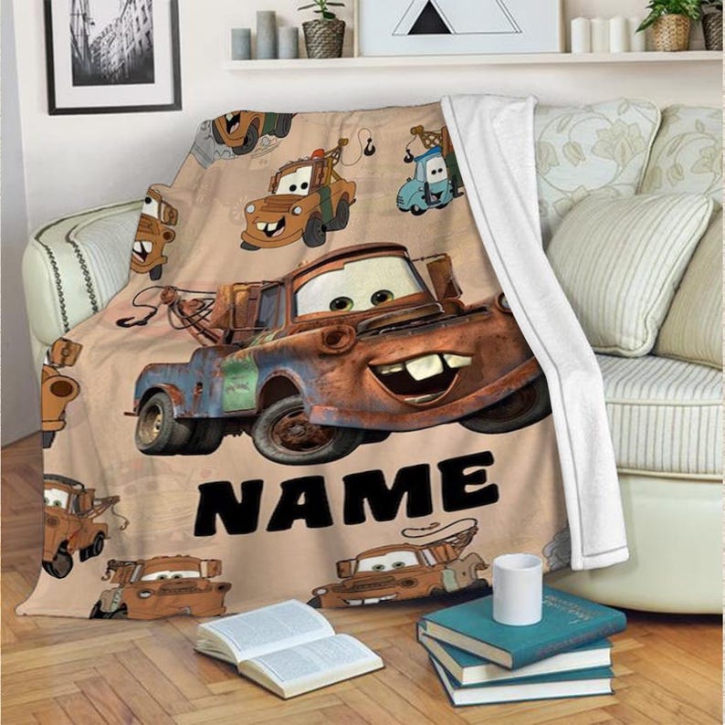 May include: A tan blanket featuring cartoon images of a rusty tow truck with a smiling face. The word "NAME" is printed in large black letters. The blanket is draped over a beige couch.