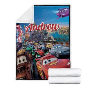 Op de afbeelding: Een zachte deken met een kleurrijk Cars-filmmotief, met de naam "Andrew". De afbeelding toont verschillende autokarakters die racen tegen een stadslandschap. Inclusief een stapel witte handdoeken.