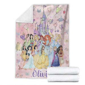 May include: Pink blanket featuring a watercolor illustration of a castle and several princesses. The blanket has a personalized name in a cursive font. Mickey Mouse-shaped balloons and butterflies decorate the background. Two folded white blankets are stacked at the bottom.