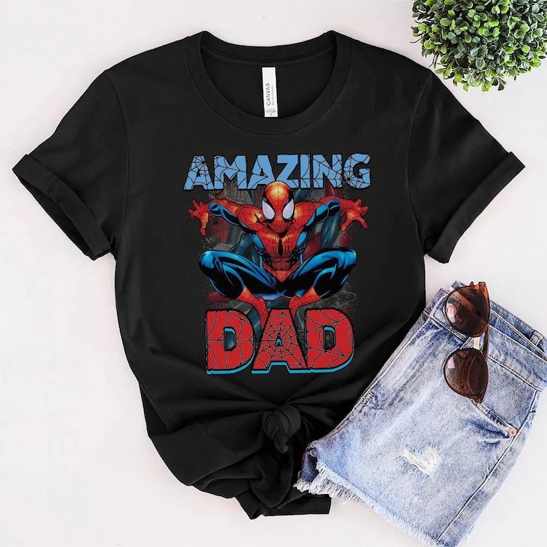 Pu&ograve; includere: Una t-shirt nera a maniche corte con la grafica di un supereroe rosso e blu in posizione accovacciata. La parola AMAZING &egrave; stampata in azzurro sopra la figura, e la parola DAD &egrave; stampata in rosso con un motivo a ragnatela sotto.