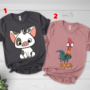 Puede incluir: Una camiseta gris oscuro con un gr&aacute;fico de un cerdito blanco con manchas marrones y una camiseta rosa empolvado con un gr&aacute;fico de un gallo colorido con ojos grandes. Ambas camisetas tienen mangas cortas, cuello redondo y una etiqueta numerada 1 y 2.