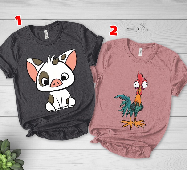 Puede incluir: Una camiseta gris oscuro con un gr&aacute;fico de un cerdito blanco con manchas marrones y una camiseta rosa empolvado con un gr&aacute;fico de un gallo colorido con ojos grandes. Ambas camisetas tienen mangas cortas, cuello redondo y una etiqueta numerada 1 y 2.