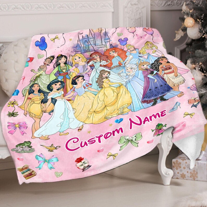 May include: Pink fleece blanket featuring a colourful illustration of Disney princesses, including Cinderella, Belle, Ariel, and Snow White. The blanket has a pink background with a castle and stars. The text "Custom Name" is printed on the blanket.