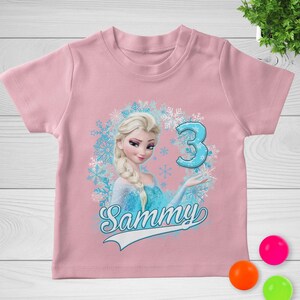 May include: A light pink short-sleeved t-shirt featuring a graphic print of a blonde character with a blue dress, surrounded by light blue snowflakes. The number 3 is printed in blue, and the name Sammy is written in a cursive script below.