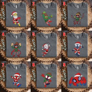 May include: A collection of nine dark gray crew neck t-shirts, each featuring a unique cartoon character dressed in holiday attire. The designs include various superheroes and villains wearing Santa hats, elf costumes, and festive red and green outfits.