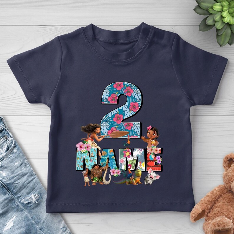 May include: A navy blue short-sleeve t-shirt featuring a large number 2 with a tropical floral pattern. Below the number, the word NAME is printed in block letters, surrounded by illustrations of Moana characters including Maui and Pua.