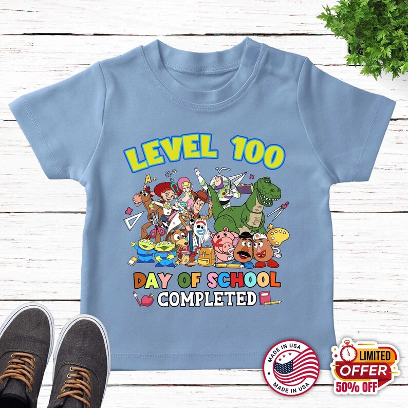 May include: A light blue t-shirt featuring a colorful graphic of Toy Story characters, including Woody, Buzz Lightyear, and Jessie. The text reads LEVEL 100 DAY OF SCHOOL COMPLETED, accompanied by school supply icons like a pencil, apple, and ruler.