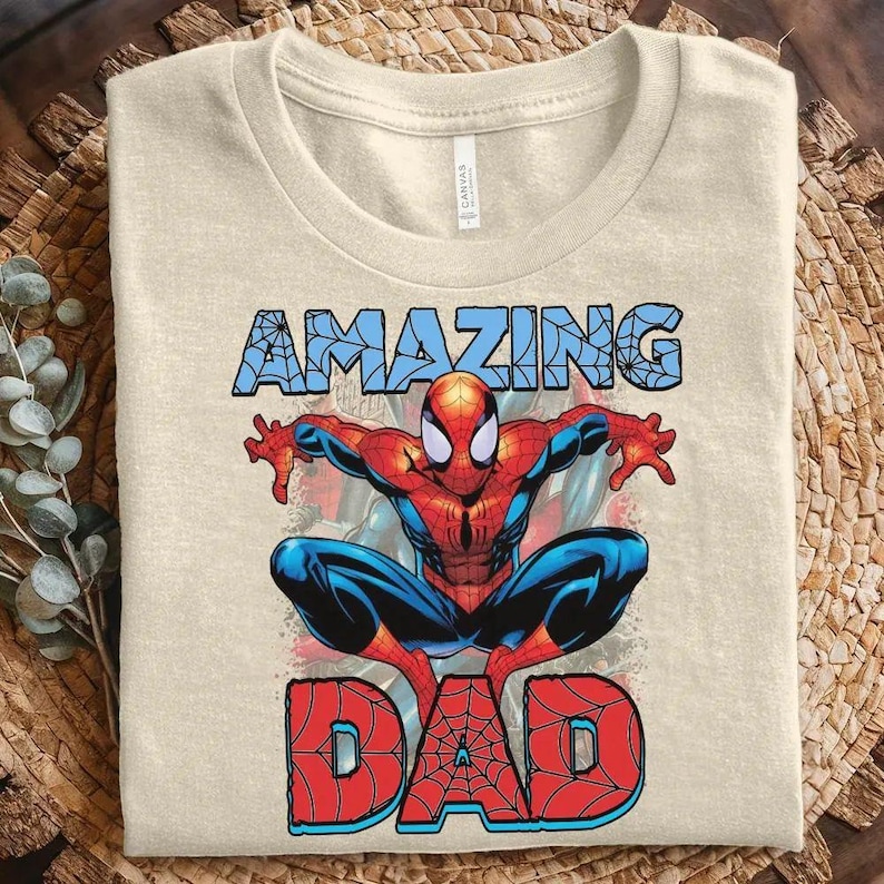 Pu&ograve; includere: Una t-shirt a girocollo color beige con una grafica del supereroe Spider-Man in posizione accovacciata. Sopra il personaggio, la parola AMAZING &egrave; stampata in azzurro con motivo a ragnatela, sotto, la parola DAD in rosso con trama a rete.