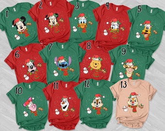 Puede incluir: Una colección de trece camisetas de manga corta en rojo, verde y rosa claro, cada una con un personaje de Disney diferente con gorro de Papá Noel y bufanda a rayas, rodeado de copos de nieve blancos, muñecos de nieve y campanas doradas.