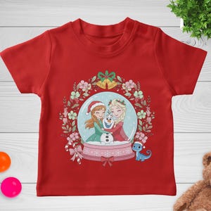 May include: A red short-sleeved t-shirt featuring a graphic of Anna, Elsa, and Olaf inside a snow globe. The globe is surrounded by a floral wreath with two gold bells at the top, a candy cane on the left, and a small blue lizard on the right.