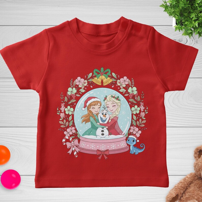 May include: A red short-sleeved t-shirt featuring a graphic of Anna, Elsa, and Olaf inside a snow globe. The globe is surrounded by a floral wreath with two gold bells at the top, a candy cane on the left, and a small blue lizard on the right.