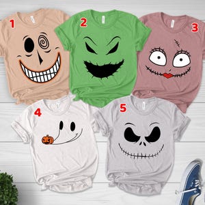 May include: Five short-sleeved t-shirts featuring character faces from The Nightmare Before Christmas. Shirt 1 is tan with a spiral eye, 2 is green with a jagged mouth, 3 is mauve with stitched eyes and lips, 4 is white with a ghost, and 5 is gray with a skull face.