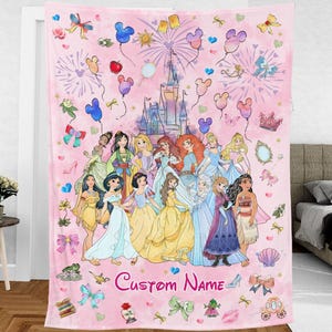 May include: A pink fleece blanket featuring a colourful illustration of Disney princesses in front of Cinderella's castle. The blanket has a pink background with hearts, stars, and other Disney-themed imagery. The text "Custom Name" is printed at the bottom of the blanket.