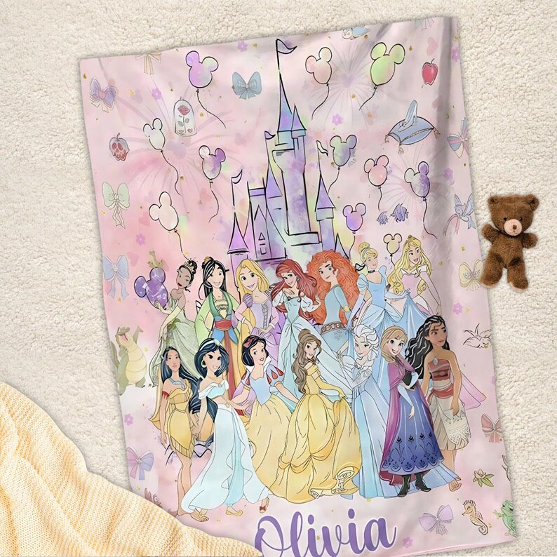 May include: A pink blanket featuring a group of Disney princesses, a castle, and Mickey Mouse-shaped balloons. The blanket has the name "Olivia" in a purple script. A small brown teddy bear is placed on the blanket.