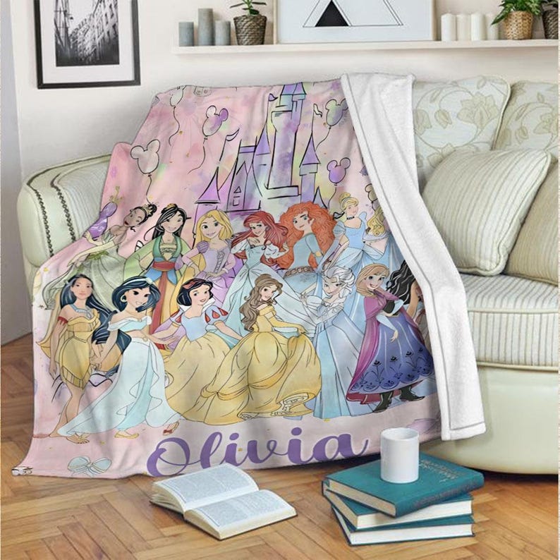 May include: A pink throw blanket featuring illustrations of various princesses, a castle, and Mickey Mouse-shaped balloons. The name "Olivia" is written in purple script across the bottom of the blanket. The blanket is draped over a sofa.