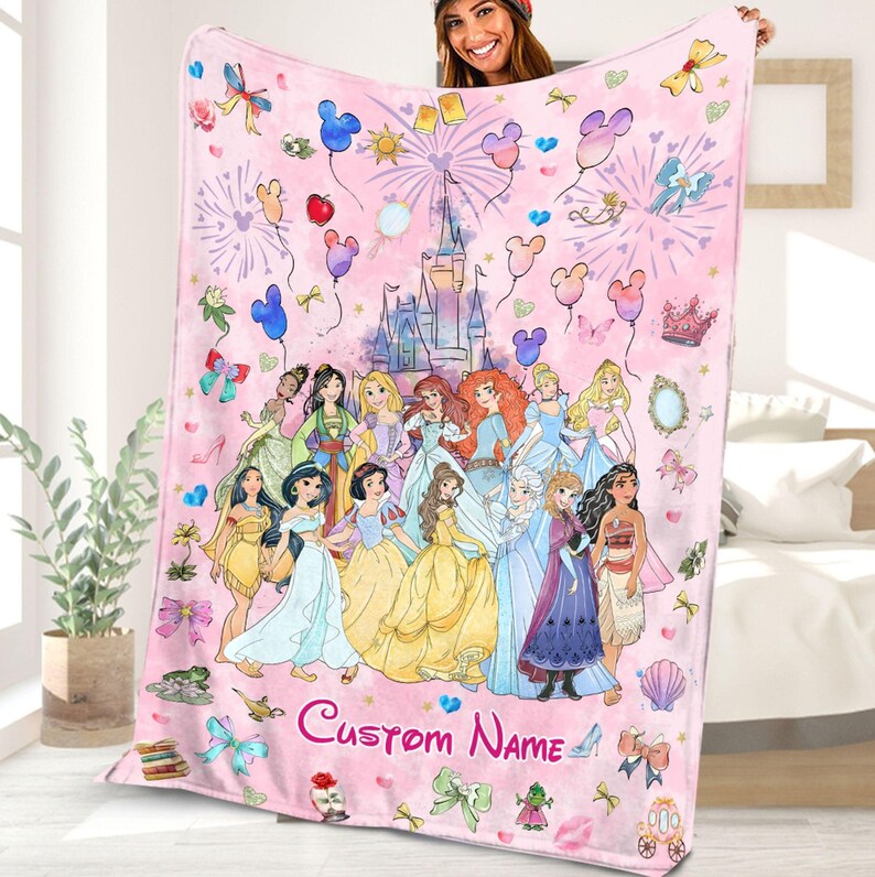 May include: Pink fleece blanket featuring a colourful illustration of Disney princesses in front of Cinderella's castle. The blanket has a pink background with hearts, stars and balloons. The text "Custom Name" is printed at the bottom of the blanket.