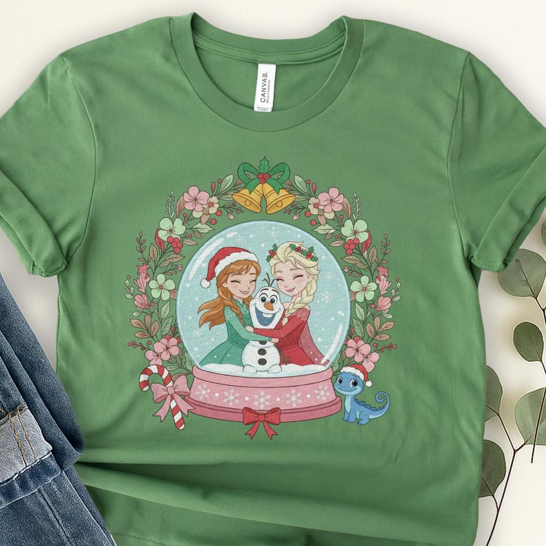 May include: A sage green t-shirt featuring a graphic of a snow globe containing cartoon characters Anna, Elsa, and Olaf. The globe is surrounded by a floral wreath with golden bells at the top, a candy cane on the left, and a small blue lizard on the right.