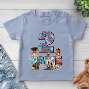 May include: A light blue short-sleeve t-shirt featuring a large number 2 with a tropical floral pattern. Below the number, the word NAME is displayed in block letters, surrounded by illustrations of Moana, Maui, Heihei, and Pua characters.