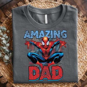 Pu&ograve; includere: Una t-shirt grigio scuro con una grafica del personaggio Spider-Man in posizione accovacciata. Sopra il personaggio c'&egrave; la parola AMAZING in azzurro con un motivo a ragnatela, e sotto la parola DAD in rosso con un motivo a ragnatela.