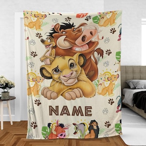 May include: A cream-colored blanket featuring characters from The Lion King. The design includes Simba, Timon, Pumbaa, and other characters, along with paw prints and the word "NAME" in brown letters. The blanket is draped over a bed.