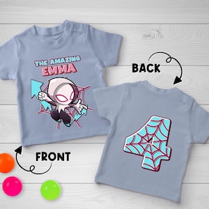May include: A light blue short-sleeve t-shirt featuring a cartoon character in a white and pink suit with the text THE AMAZING EMMA on the front, and a large number 4 with a blue and red spiderweb pattern on the back of the shirt.
