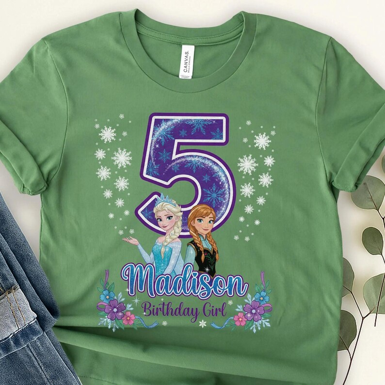 以下が含まれることがあります： セージグリーンのTシャツ。雪の結晶で飾られた大きな紫色の数字の5が中央にあります。数字の下には、2人のアニメキャラクターのイラスト、Madisonという名前、Birthday Girlという文字が描かれ、カラフルな花のアクセントが添えられています。