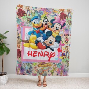 May include: A colorful blanket featuring Disney characters Mickey Mouse, Minnie Mouse, Donald Duck, and Goofy. The blanket has a pink border with the name "HENRY" in red. The background is filled with various Disney character illustrations.