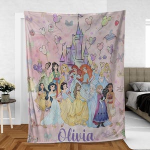 May include: A pink blanket featuring a group of cartoon princesses in colorful gowns, with a castle and balloons in the background. The name "Olivia" is written in purple script at the bottom.