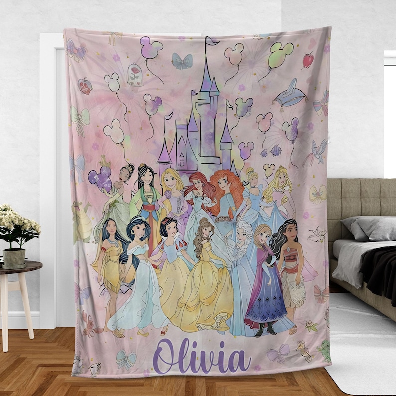 May include: A pink blanket featuring a group of cartoon princesses in colorful gowns, with a castle and balloons in the background. The name "Olivia" is written in purple script at the bottom.