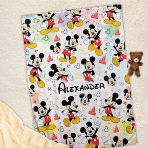 K&ouml;nnte beinhalten: Eine wei&szlig;e Decke mit einem sich wiederholenden Muster von Mickey Mouse, roten Burgen, Herzen und Sternen. Der Name "Alexander" ist in schwarzer Schrift gedruckt. Ein kleiner brauner Teddyb&auml;r liegt daneben.