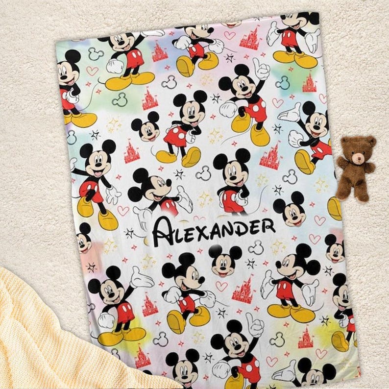 K&ouml;nnte beinhalten: Eine wei&szlig;e Decke mit einem sich wiederholenden Muster von Mickey Mouse, roten Burgen, Herzen und Sternen. Der Name "Alexander" ist in schwarzer Schrift gedruckt. Ein kleiner brauner Teddyb&auml;r liegt daneben.