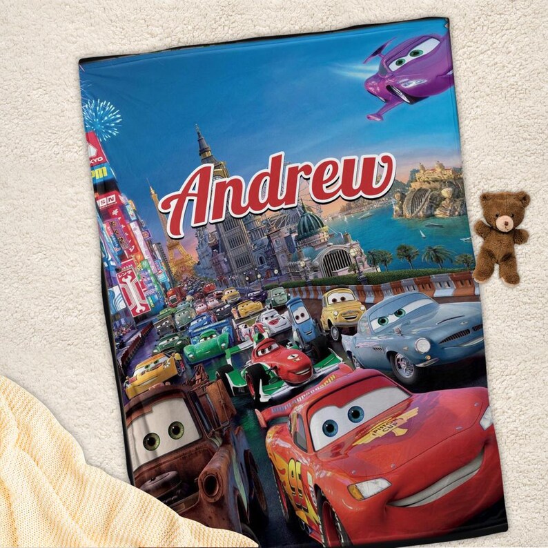 Op de afbeelding: Een gepersonaliseerde deken met een sc&egrave;ne uit de film Cars, met de naam "Andrew". Het ontwerp toont verschillende autokarakters die racen tegen een stadsgezicht. Levendige kleuren.