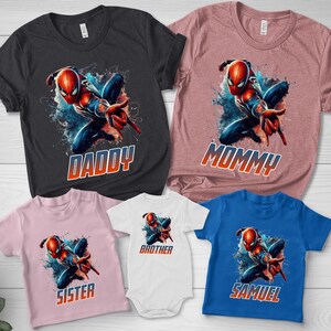 May include: A collection of five t-shirts and one bodysuit featuring a graphic of a red and blue superhero in a dynamic pose. The shirts are black, mauve, light pink, white, and blue, with the text DADDY, MOMMY, SISTER, BROTHER, and SAMUEL.